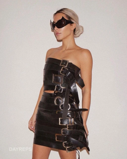 A woman poses in a black, buckled two-piece outfit, accessorized with sunglasses, against a neutral backdrop, creating a high-fashion studio look.