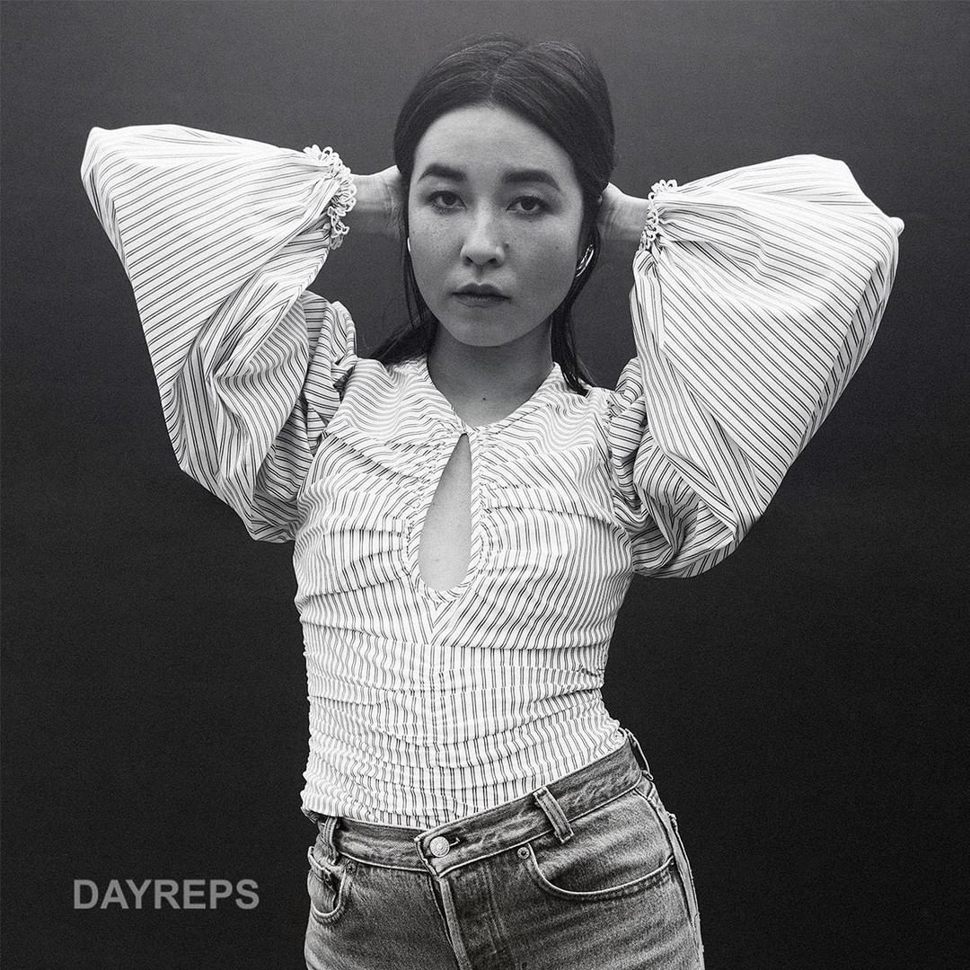 Monochrome portrait of a young Asian woman with hands behind her head, wearing a striped top and jeans against a dark background.