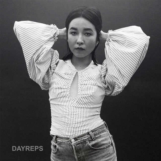 Monochrome portrait of a young Asian woman with hands behind her head, wearing a striped top and jeans against a dark background.