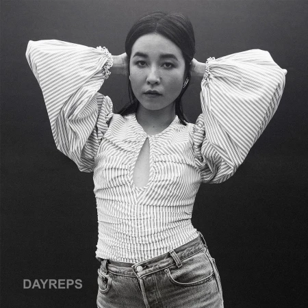 Monochrome portrait of a young Asian woman with hands behind her head, wearing a striped top and jeans against a dark background.