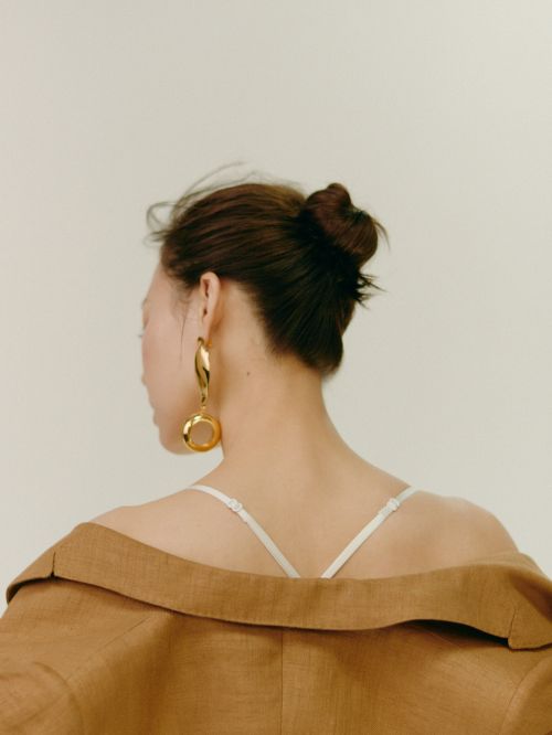 Brunette woman poses in tan jacket and large gold earrings against a white background in a studio shot for a beauty fashion editorial.