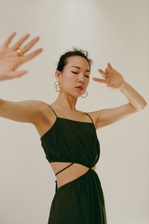 An Asian woman in a cut-out black dress poses against a neutral background, wearing hoop earrings and a ring while facing forward.