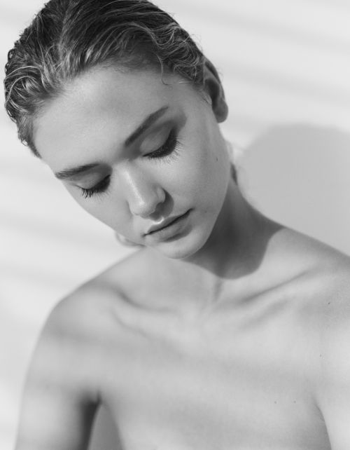 Black and white portrait of a young woman with wet hair looking down in a photo studio with striped shadow pattern in the background.