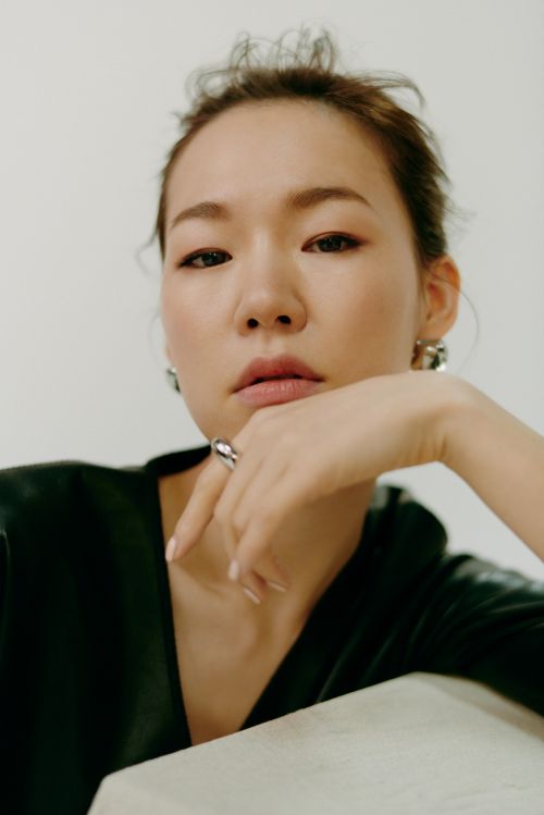 Close-up portrait of an Asian woman with dark hair, wearing a black top and silver jewelry, resting her chin on her hand indoors.