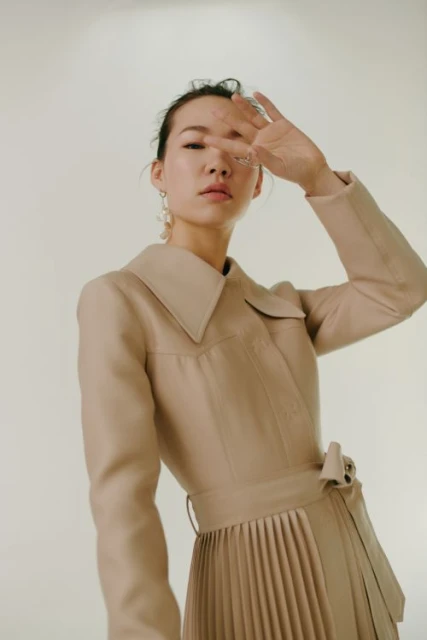 Asian woman poses in a beige dress with a pleated skirt, under bright lighting in a studio. Hand is raised, partially obscuring her face.