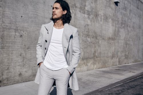 Man in grey coat, white shirt, and grey pants stands against a concrete wall. Fashion shot in natural light, gazing to the left.