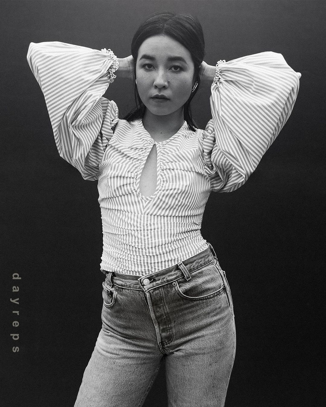 Woman in a striped blouse and jeans poses against a dark background, hands behind her head, looking directly at the camera.