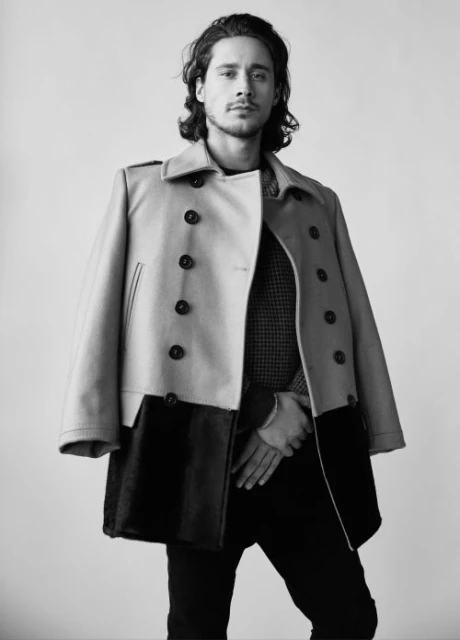 Portrait of a man in a tan overcoat with a black skirt; he is wearing dark pants and has medium-length curly brown hair.