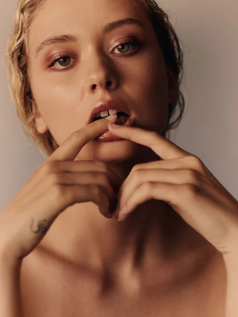 Close-up portrait of a young adult woman with pink eye shadow, her fingers near her mouth, set against a neutral studio background.