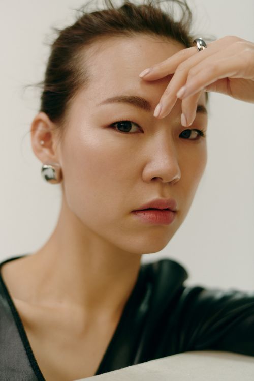 Close-up of an Asian woman with dark hair styled back, wearing a dark top, silver earrings and a ring. She is posing indoors.