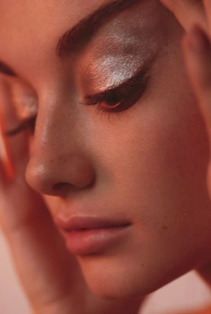 Close-up beauty shot of a woman with iridescent eyeshadow, peachy skin tone, and soft, diffused lighting in an indoor setting.