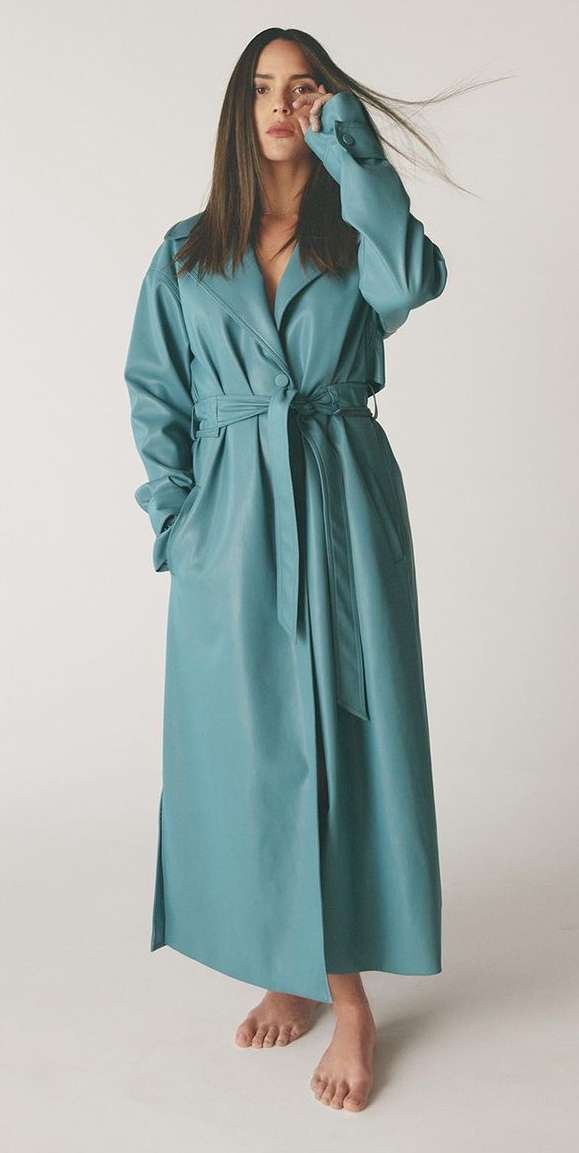 A woman with long brown hair poses in a studio wearing a long blue trench coat cinched at the waist, striking a fashion-forward pose.