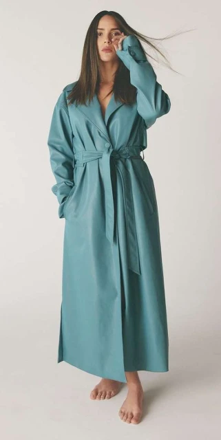 A woman with long brown hair poses in a studio wearing a long blue trench coat cinched at the waist, striking a fashion-forward pose.
