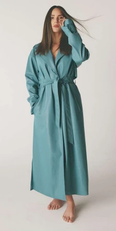 A woman with long brown hair poses in a studio wearing a long blue trench coat cinched at the waist, striking a fashion-forward pose.