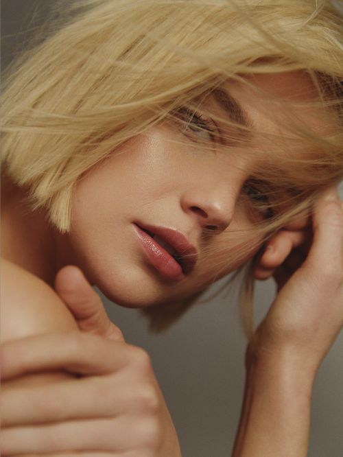 Close-up portrait of a woman with short blonde hair and parted lips, framed by loose strands blowing across her face.
