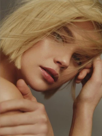 Close-up portrait of a woman with short blonde hair and parted lips, framed by loose strands blowing across her face.