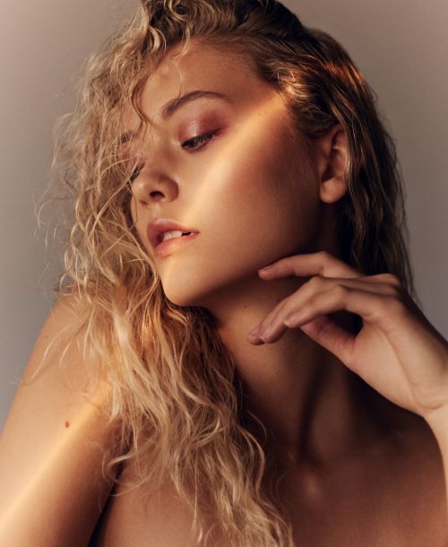 Close-up studio portrait of a young adult woman with blonde curly hair and pink eye shadow, gazing downward in soft, diffused light.