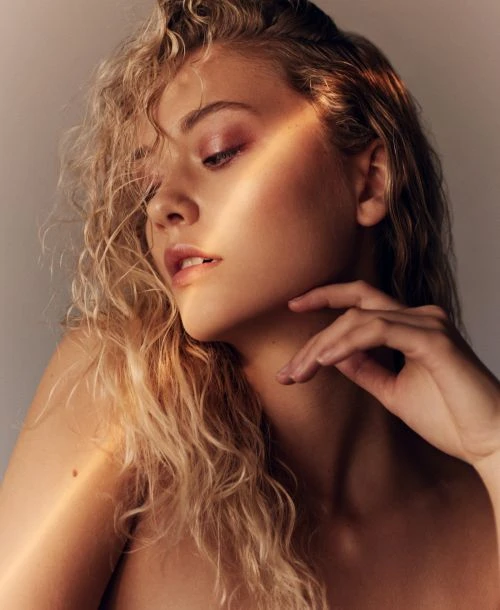 Close-up studio portrait of a young adult woman with blonde curly hair and pink eye shadow, gazing downward in soft, diffused light.