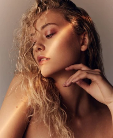 Close-up studio portrait of a young adult woman with blonde curly hair and pink eye shadow, gazing downward in soft, diffused light.
