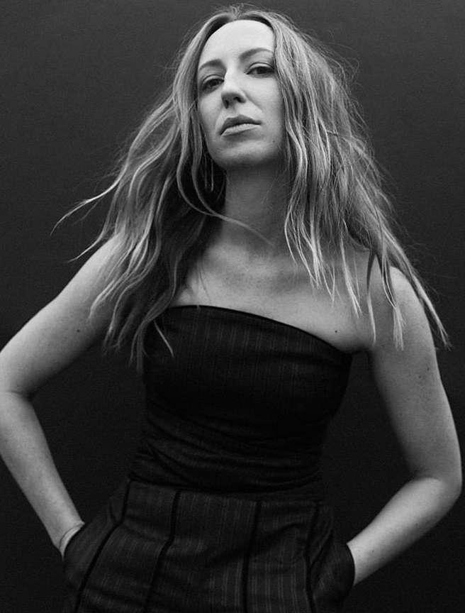 Black and white portrait of an adult woman with long blonde hair, wearing a dark tube top, hands on hips, against a dark backdrop.