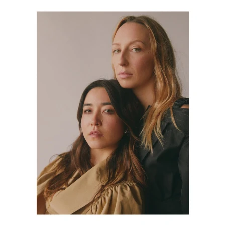 Portrait of two women together, one in a tan collared garment and one in a dark top against a light grey background