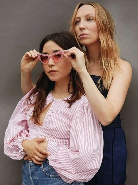 Two adult women pose indoors against a gray wall. One wears pink heart-shaped sunglasses and a pink striped shirt. The other wears a strapless dress.