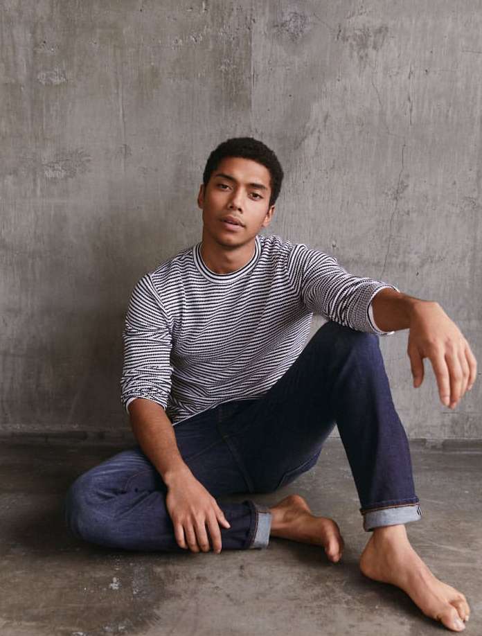Young man sitting barefoot on the floor in jeans and a striped shirt, looking at the camera, with a concrete wall background.