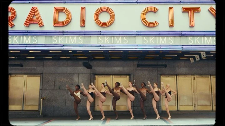 A line of dancers in leotards perform high kicks outside Radio City Music Hall beneath the SKIMS advertisement sign.
