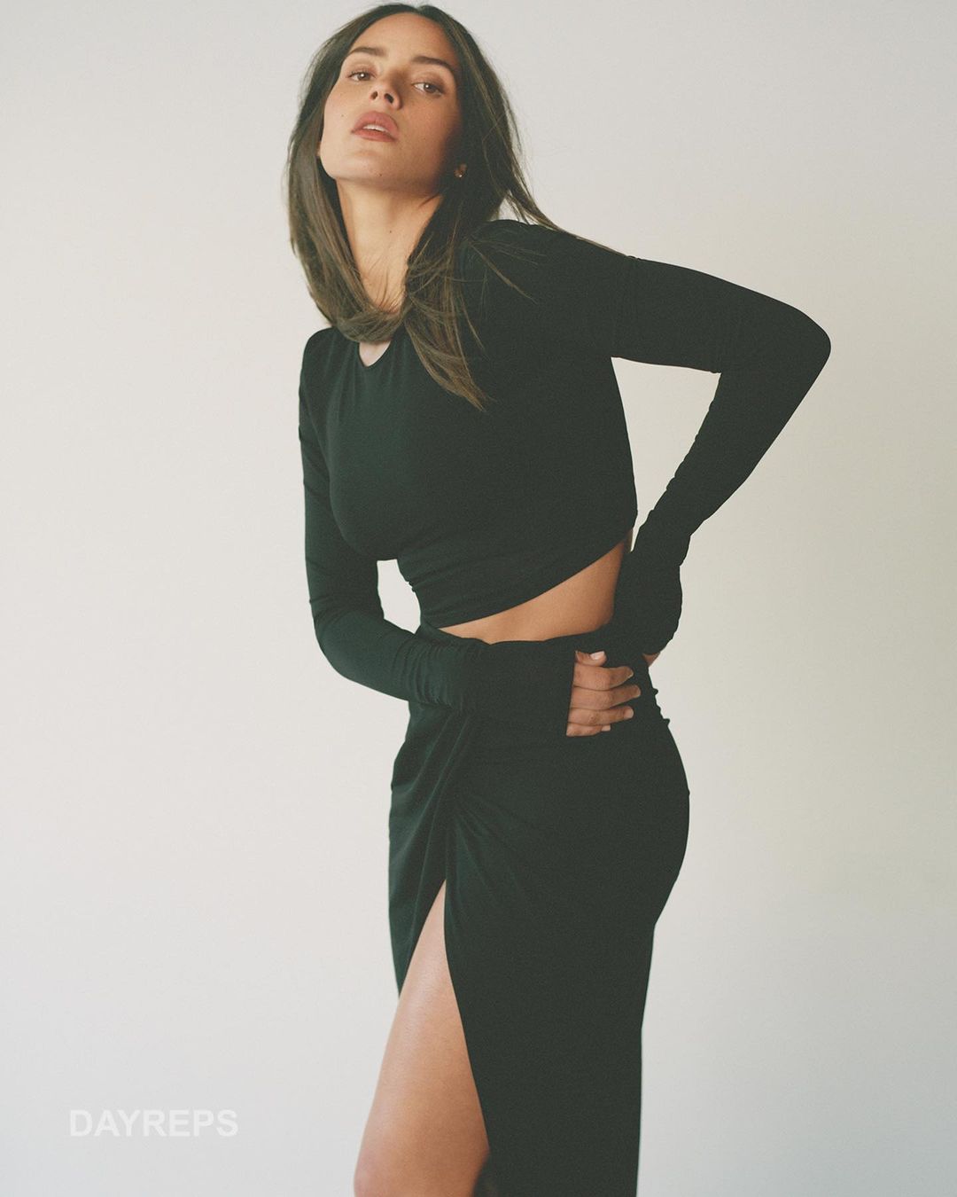 Woman wearing a black long-sleeve crop top and matching midi skirt with a high slit against a plain white studio background.