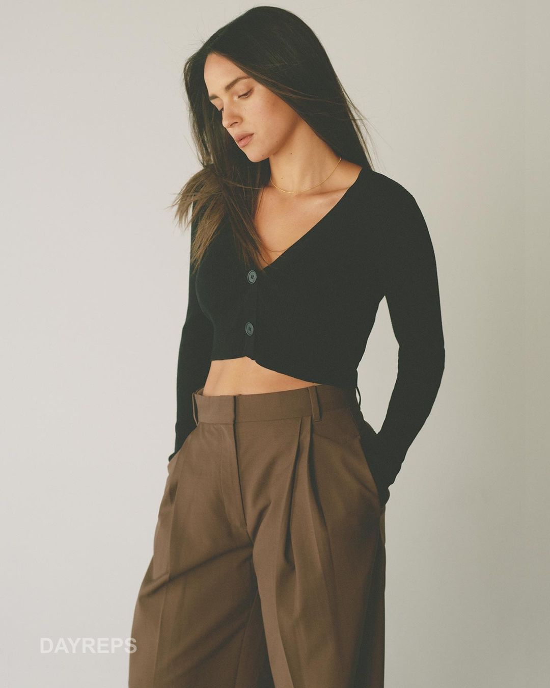 Woman in black V-neck cardigan and brown pleated trousers posing against a plain neutral background by photographer Gray Hamner.