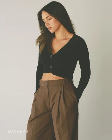 Woman in black V-neck cardigan and brown pleated trousers posing against a plain neutral background by photographer Gray Hamner.