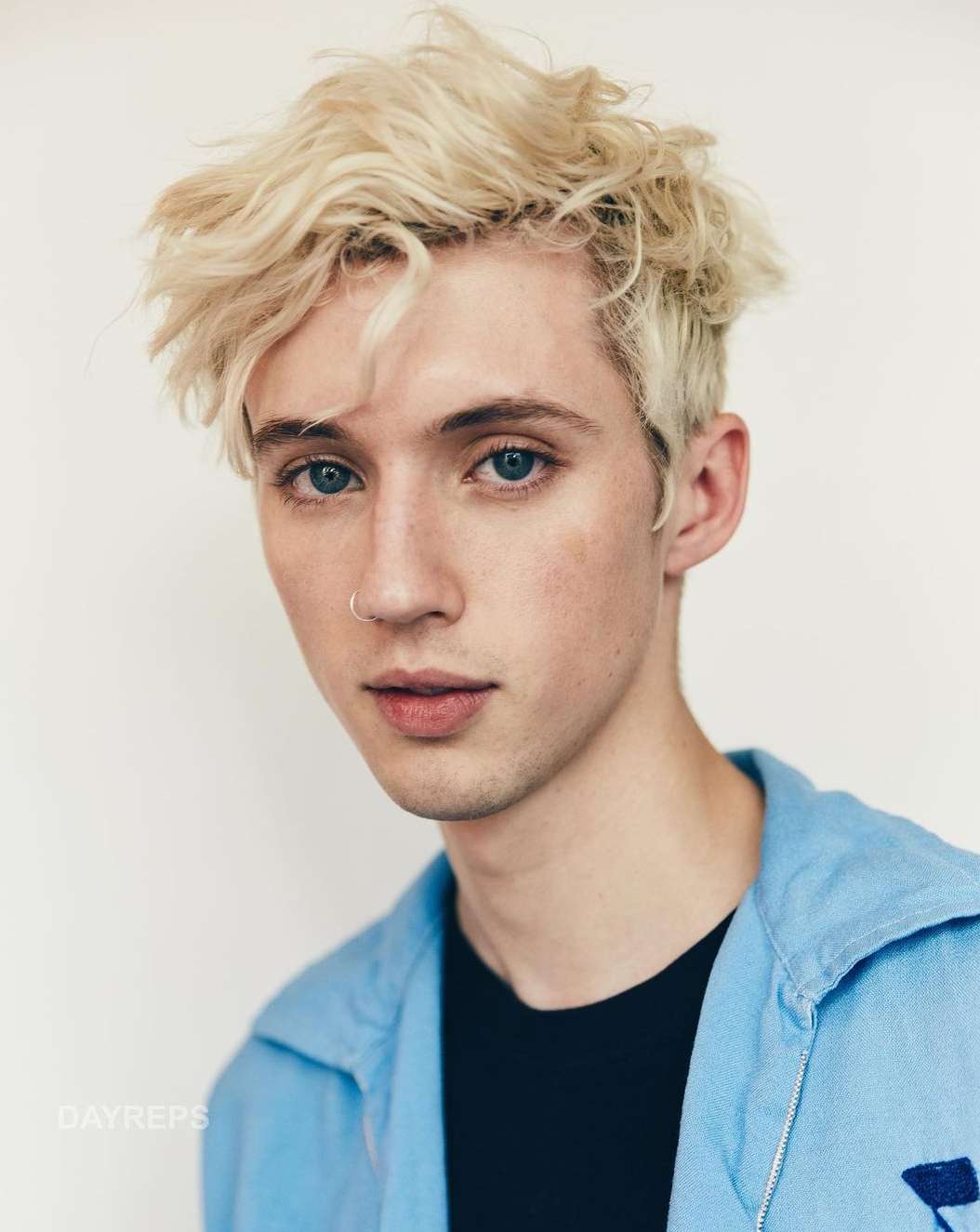 Close-up portrait of a blonde male with light skin and blue eyes, wearing a blue shirt and a small nose ring, against a white background.