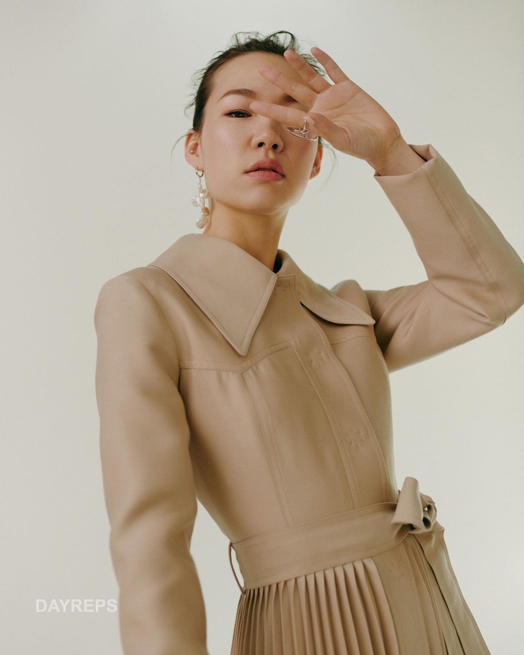 A low-angle shot features a stylish woman in a trench coat with pleated skirt, covering her face with her hand in a fashion pose.