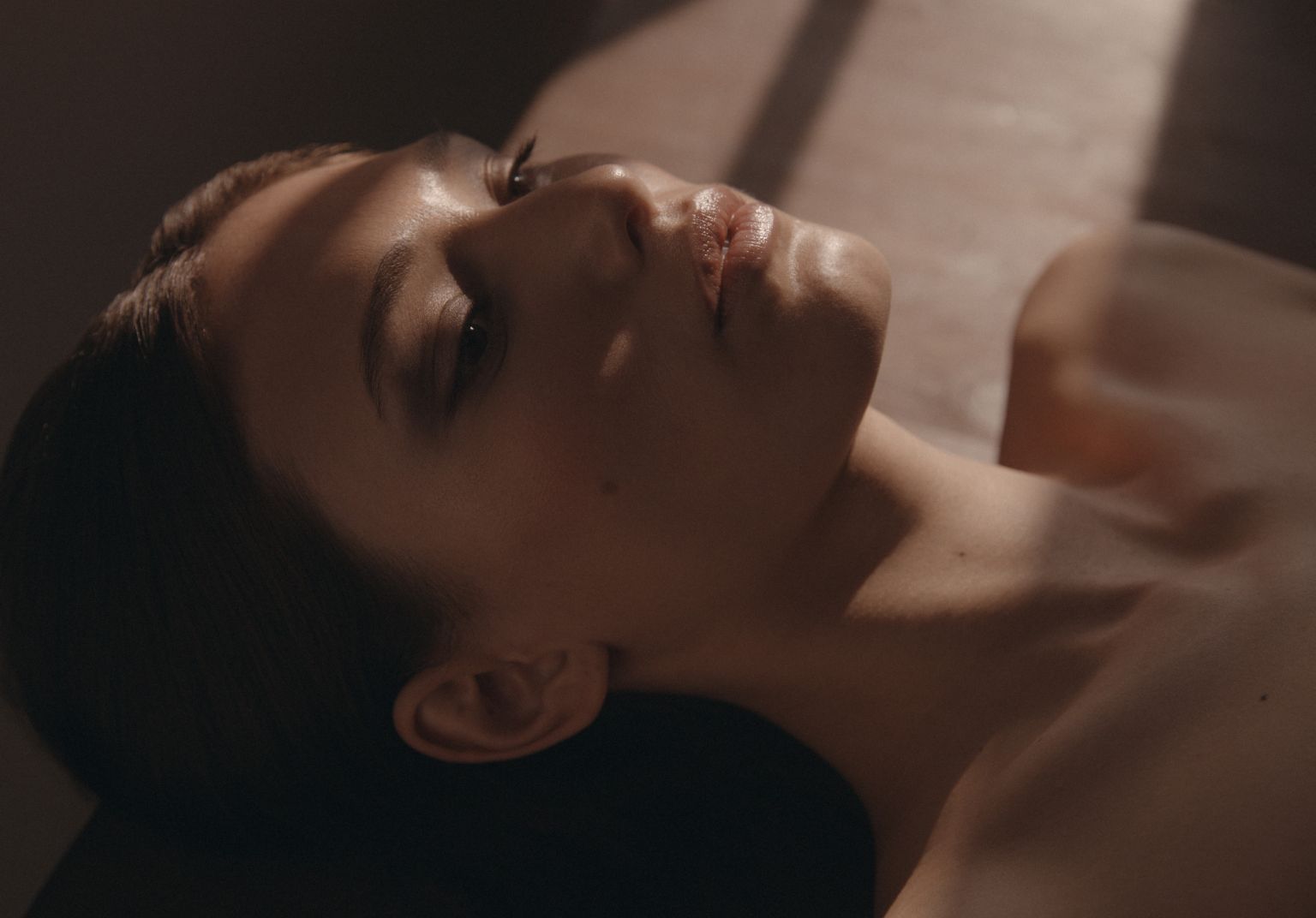 Close-up studio shot of an adult female lying on her back with her eyes open, showcasing her face and upper chest under warm, soft lighting.