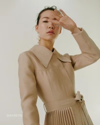 Model in a beige leather coat with a pleated skirt, holding a hand up near her face against a plain background