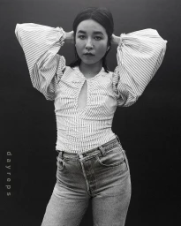 Portrait of a woman wearing a striped puff-sleeve top and jeans with hands behind her head against a dark backdrop