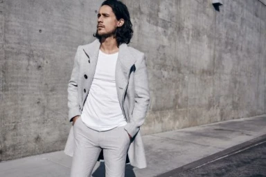 A man with long dark hair wearing a light grey overcoat and white t-shirt standing outdoors against a concrete wall.