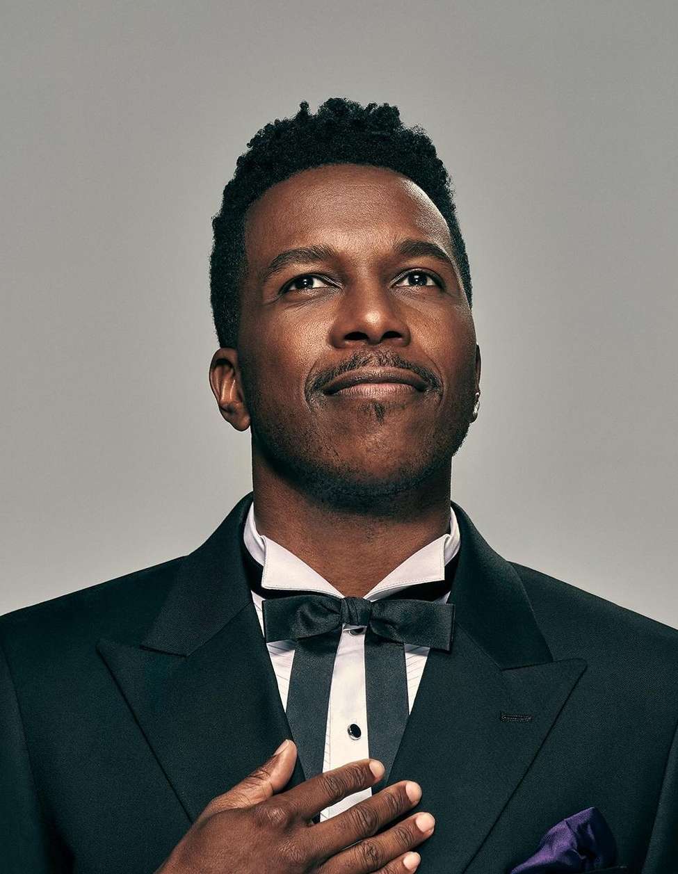 Portrait of a black man wearing a black tuxedo and bow tie looking upward against a plain light grey studio backdrop.