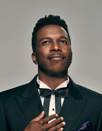 Portrait of a black man wearing a black tuxedo and bow tie looking upward against a plain light grey studio backdrop.