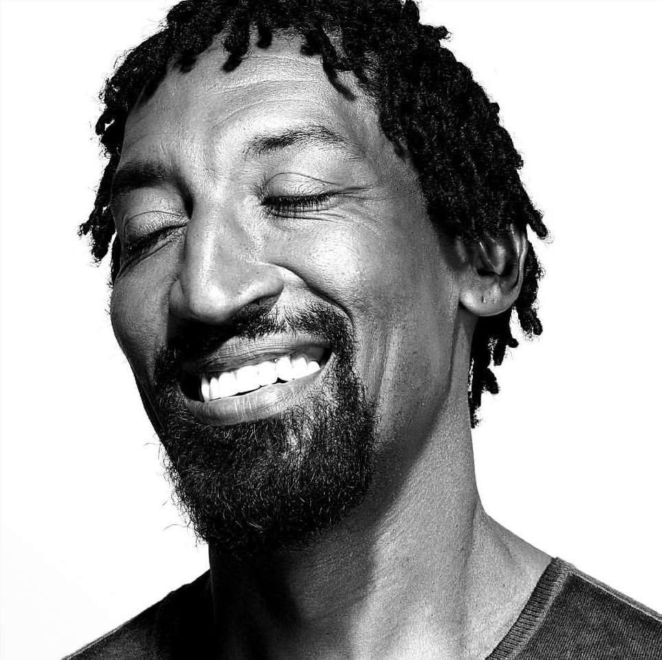 Black and white close-up portrait of a smiling man with dreadlocks, eyes closed, and a beard against a plain background.