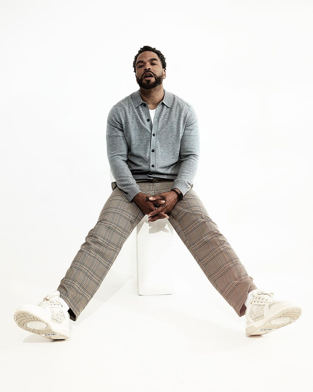 Adult man with a beard sits with legs spread in grey cardigan, light-brown plaid pants, and sneakers against a plain white backdrop.