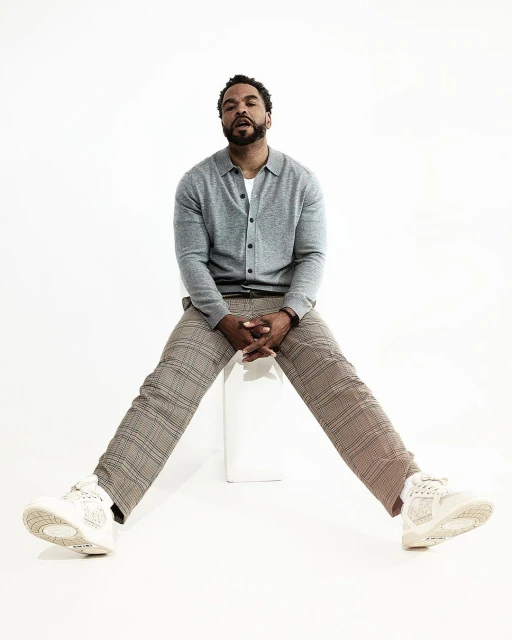 Adult man with a beard sits with legs spread in grey cardigan, light-brown plaid pants, and sneakers against a plain white backdrop.