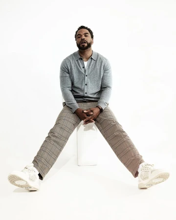 Adult man with a beard sits with legs spread in grey cardigan, light-brown plaid pants, and sneakers against a plain white backdrop.