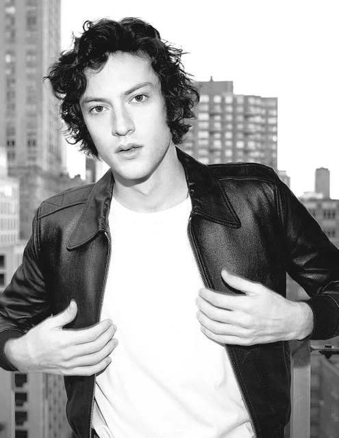 Monochrome portrait of a young adult male with curly hair in a leather jacket standing against an urban cityscape backdrop.
