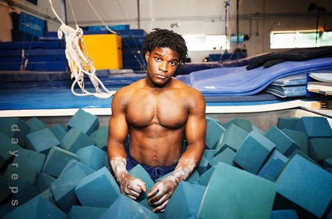 Athletic man with muscular build is surrounded by foam blocks in an indoor training gym; shirtless with a serious facial expression.