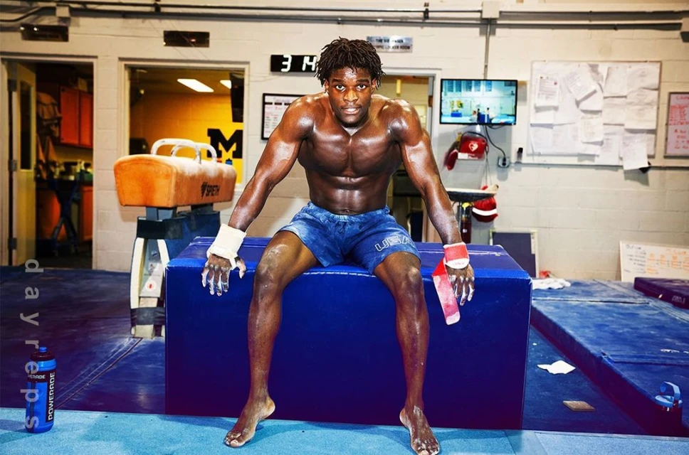 Muscular man with dark skin seated in a gym with training equipment, wearing blue shorts and wrist wraps, gazing at the camera.