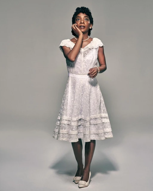 Woman in vintage dress and heels poses in a studio with hand to face, looking upwards in portrait orientation with a neutral background.