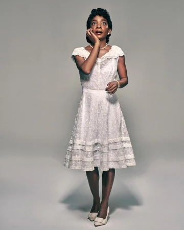 Woman in vintage dress and heels poses in a studio with hand to face, looking upwards in portrait orientation with a neutral background.