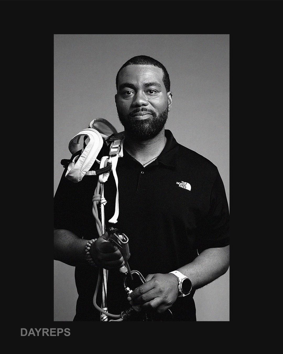 Monochromatic portrait of a Black adult male in a polo shirt holding climbing gear, with climbing harness over his shoulder.