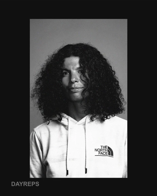 Black and white portrait of a man with curly hair wearing a The North Face hooded sweatshirt, captured in an indoor photo studio.